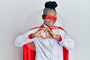 Young african american woman wearing doctor uniform and super hero costume smiling in love doing heart symbol shape with hands