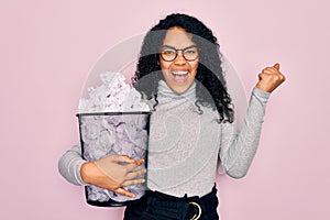 Young african american woman wearing glasses holding wastebasket with cumpled papers screaming proud and celebrating victory and