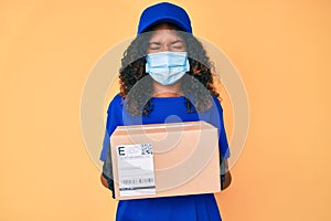 Young african american woman wearing medical mask holding delivery package sticking tongue out happy with funny expression