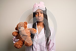 Young african american woman wearing pajama and eye mask hugging teddy bear with a confident expression on smart face thinking