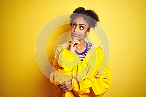 Young african american woman wearing rain coat over isolated yellow background with hand on chin thinking about question, pensive