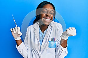 Young african american woman wearing scientist uniform holding syringe screaming proud, celebrating victory and success very