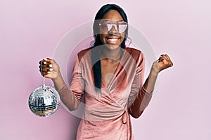 Young african american woman wearing sexy party dress holding disco ball screaming proud, celebrating victory and success very