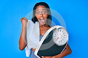 Young african american woman wearing sportswear holding weighing machine screaming proud, celebrating victory and success very