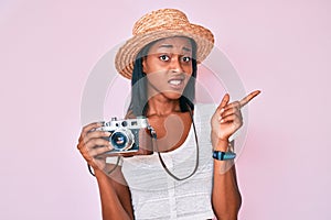 Young african american woman wearing summer hat holding vintage camera pointing aside worried and nervous with forefinger,