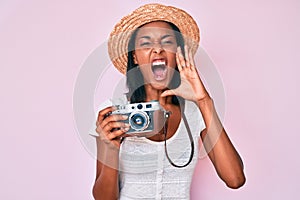 Young african american woman wearing summer hat holding vintage camera shouting and screaming loud to side with hand on mouth
