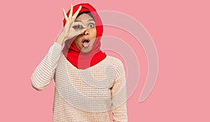 Young african american woman wearing traditional islamic hijab scarf doing ok gesture shocked with surprised face, eye looking