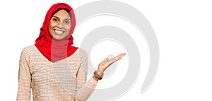 Young african american woman wearing traditional islamic hijab scarf smiling cheerful presenting and pointing with palm of hand