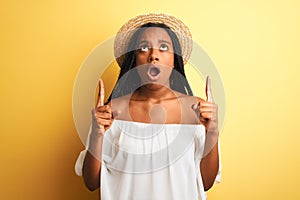 Young african american woman wearing white t-shirt and hat over  yellow background amazed and surprised looking up and