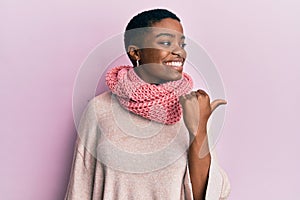 Young african american woman wearing winter scarf smiling with happy face looking and pointing to the side with thumb up