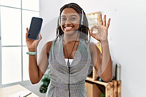 Young african american woman working at the office wearing operator headset holding smartphone doing ok sign with fingers, smiling