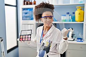 Young african american woman working at scientist laboratory doing make up pointing thumb up to the side smiling happy with open