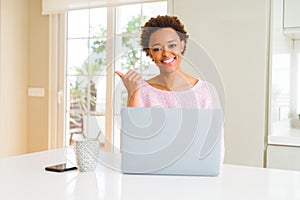 Young african american woman working using computer laptop smiling with happy face looking and pointing to the side with thumb up