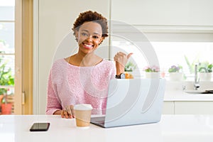 Young african american woman working using computer laptop smiling with happy face looking and pointing to the side with thumb up