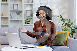 A young African-American woman works at home, sits on the couch in a headset, holds a tablet in her hands, and talks on