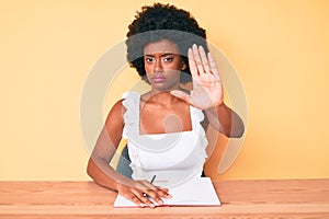 Young african american woman writing book with open hand doing stop sign with serious and confident expression, defense gesture