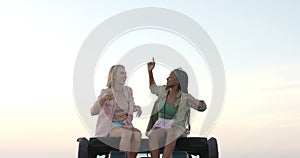 Young African American woman and young Caucasian woman sit atop a vehicle outdoors on a road trip