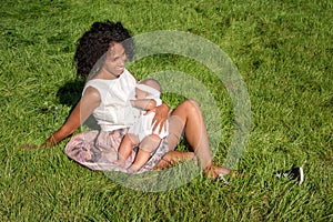 Young african american woman breastfeeding her baby sitting on grass in summer park. Infancy, motherhood, lactation, nutrition and