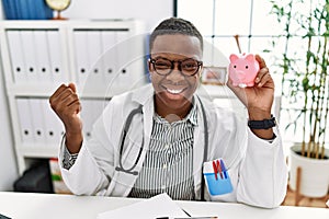 Young african doctor man holding piggy bank at the clinic screaming proud, celebrating victory and success very excited with