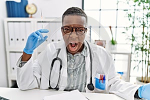 Young african doctor man holding syringe at the hospital angry and mad screaming frustrated and furious, shouting with anger