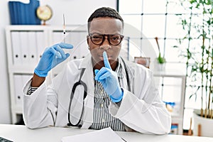 Young african doctor man holding syringe at the hospital asking to be quiet with finger on lips