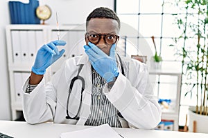 Young african doctor man holding syringe at the hospital shocked covering mouth with hands for mistake