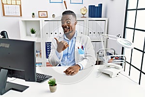 Young african doctor man wearing stethoscope at the clinic smiling with happy face looking and pointing to the side with thumb up