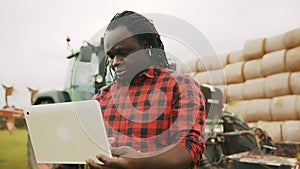 Young african farmer using tablet in front of big green tractor anf haystack