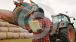 Young african farmer using tablet in front of big green tractor anf haystack