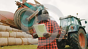 Young african farmer using tablet in front of big green tractor anf haystack