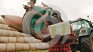 Young african farmer using tablet in front og big green tractor anf haystack