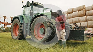 Young african farmer using tablet in front og big green tractor anf haystack