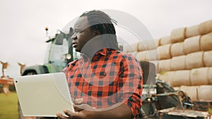 Young african farmer using tablet in front og big green tractor and haystack