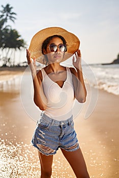 Young african female model in straw hat and sunglasses posing at resort by sea at sunrise. Black woman against scenic