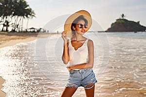 Young african female model in straw hat and sunglasses posing at resort by sea at sunrise. Black woman against scenic
