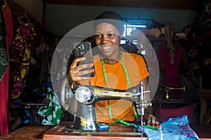 Young african female tailor smiling while viewing content on her mobile phone sitting with her sewing machine.