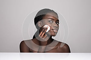 Young african girl applying foundation on her face using makeup sponge. Portrait of smiling african girl on white background