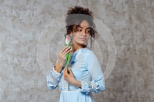 Young African girl in a blue dress with a Tulip on a gray background. The concept of women`s day and spring