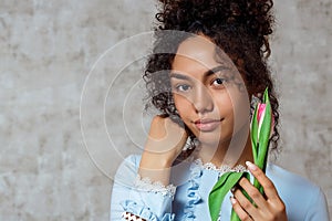 Young African girl in a blue dress with a Tulip on a gray background. The concept of women`s day and spring