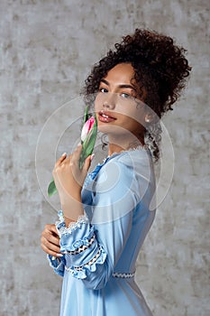 Young African girl in a blue dress with a Tulip on a gray background. The concept of women`s day and spring