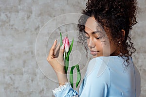 Young African girl in a blue dress with a Tulip on a gray background. The concept of women`s day and spring