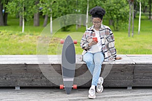 Young african girl chat using smartphone application sitting outdoors with longboard skate in park