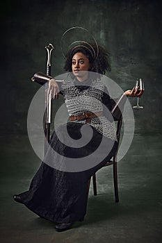 Young African girl, medieval warrior in chainmail armor sitting on chair with sword and wine glass against vintage green