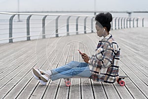 Young african girl sit on longboard with smartphone in hand text outdoor in park using 5g connection
