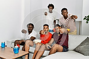 Young african group of friends sitting on the sofa at home playing video games pointing with finger to the camera and to you,