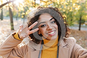 Young african happy woman walking outdoors in a spring park take a selfie by camera make peace gesture