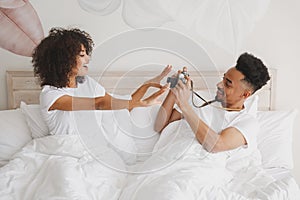 Young african loving couple girl guy in white t-shirts lying on bed doing shot on retro vintage photo camera in bedroom