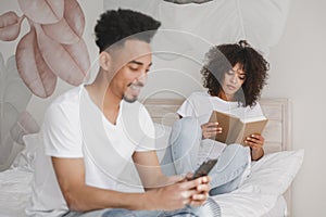 Young african loving couple girl guy in white t-shirts reading book using mobile phone lying on bed indoors in bedroom