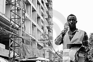 Young African man construction worker thinking while talking on the phone at building site