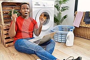 Young african man doing laundry and using computer amazed and surprised looking up and pointing with fingers and raised arms